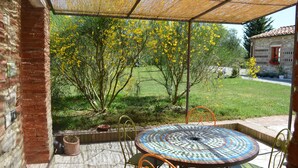 A covered patio with a mosaic table and chairs, a brick wall, and a view of a garden with yellow flowering trees. - Apartment With Beautiful View in the Crete Senesi (Asciano)