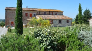A two-story stone building with a tiled roof, surrounded by lush greenery and tall trees. - Apartment With Beautiful View in the Crete Senesi (Asciano)