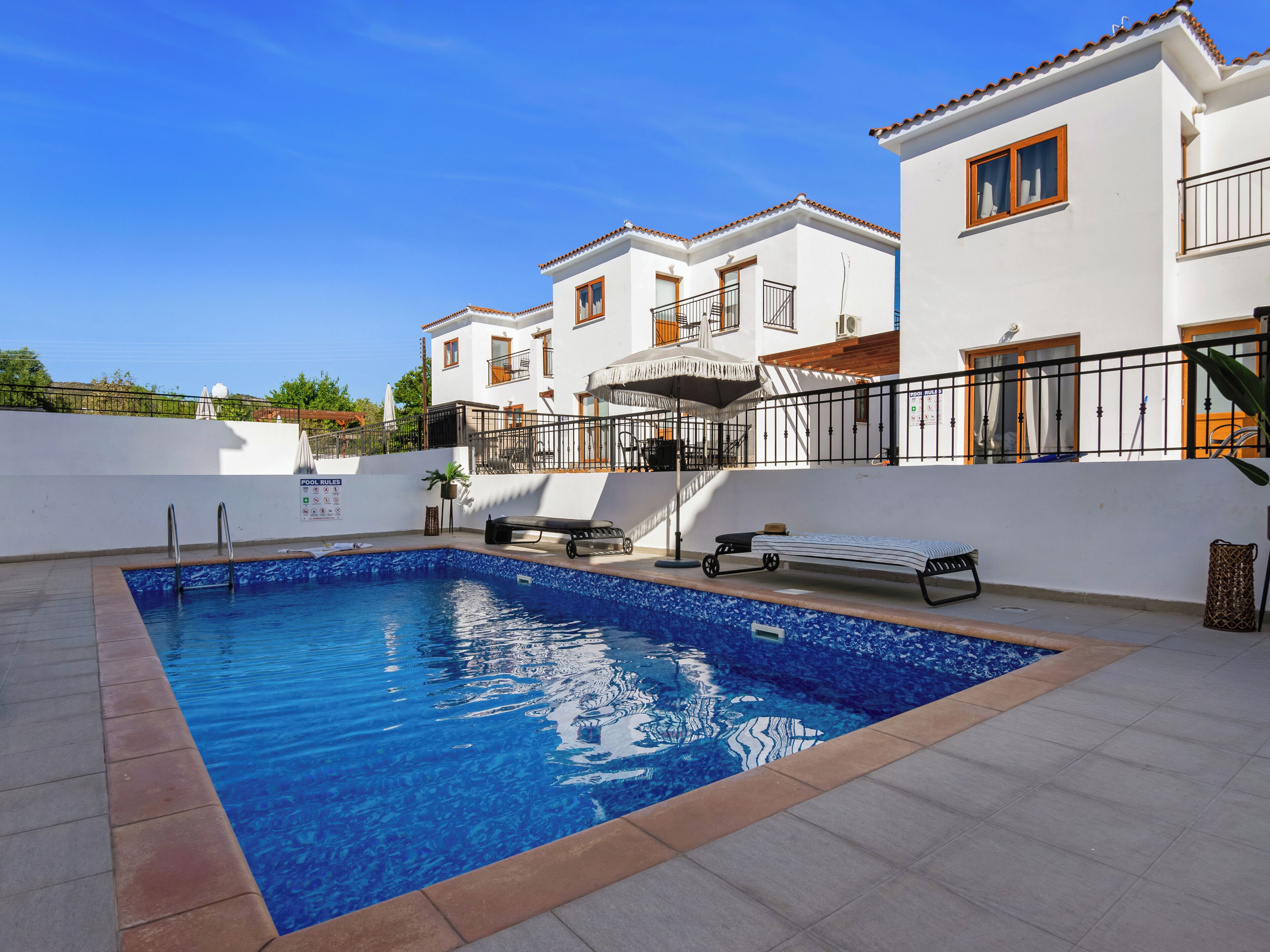 A swimming pool with a tiled deck, a lounge area with a sunbed, and a white building with balconies.
