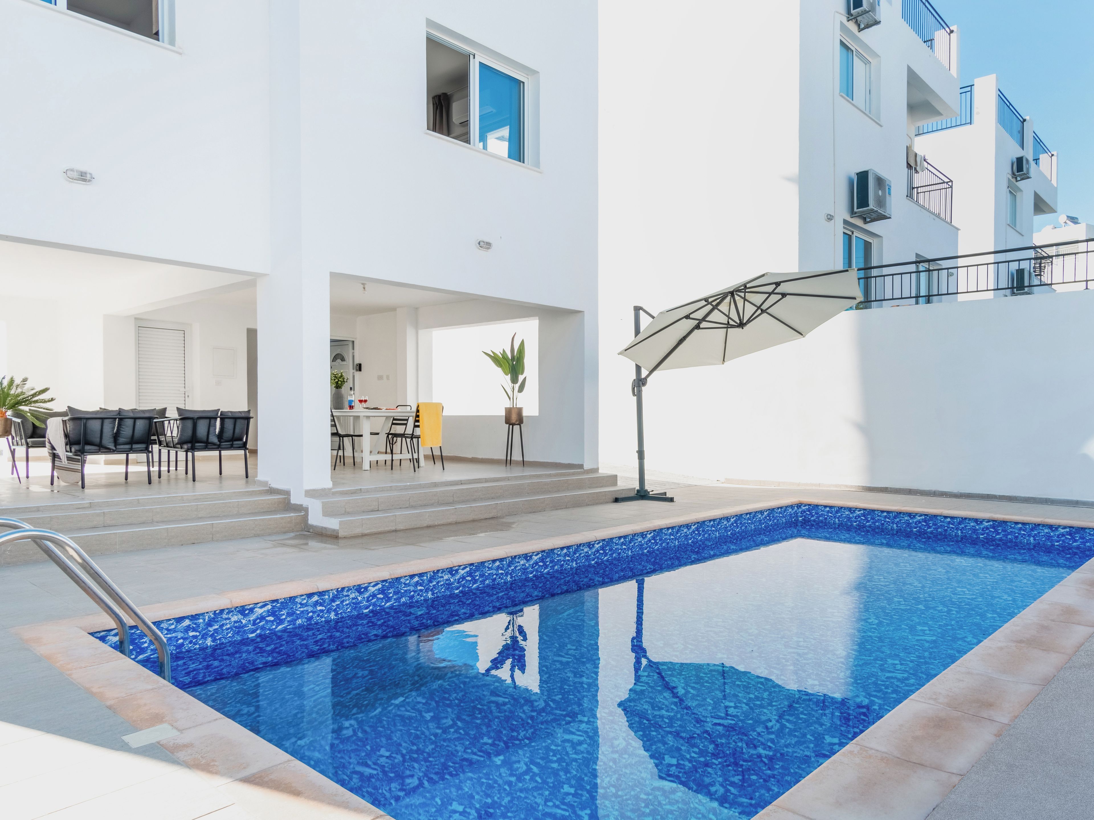 A modern outdoor pool area with a clear blue pool, a dining area with chairs and a table, and a staircase leading to an upper level.