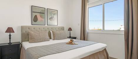 A bedroom with a bed, two bedside tables, a lamp, a picture frame, an air conditioning unit, and a window with curtains.