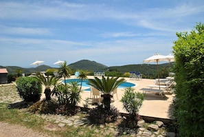 A pool area with lounge chairs and umbrellas, surrounded by greenery and hills in the background.