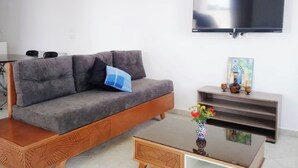 A living room with a sofa, coffee table, TV, and a small shelf with decorative items.