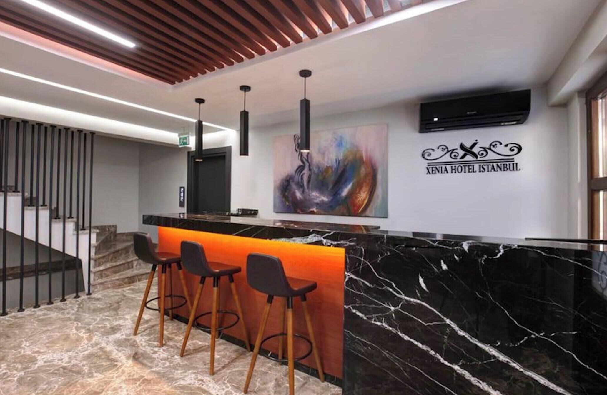 Un espace d’accueil d’hôtel avec un comptoir en marbre, trois tabourets de bar, un design moderne au plafond et un téléviseur fixé au mur.