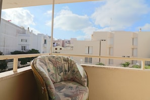 Een balkon met een rieten stoel, uitzicht op woongebouwen en een heldere lucht.
