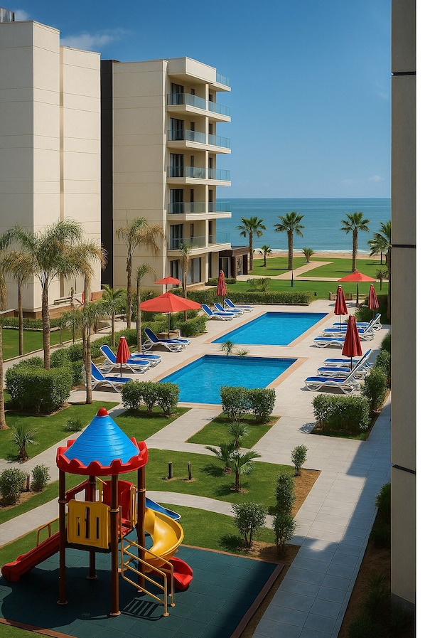 A resort pool area with a children's play structure, lounge chairs, and a view of the ocean.