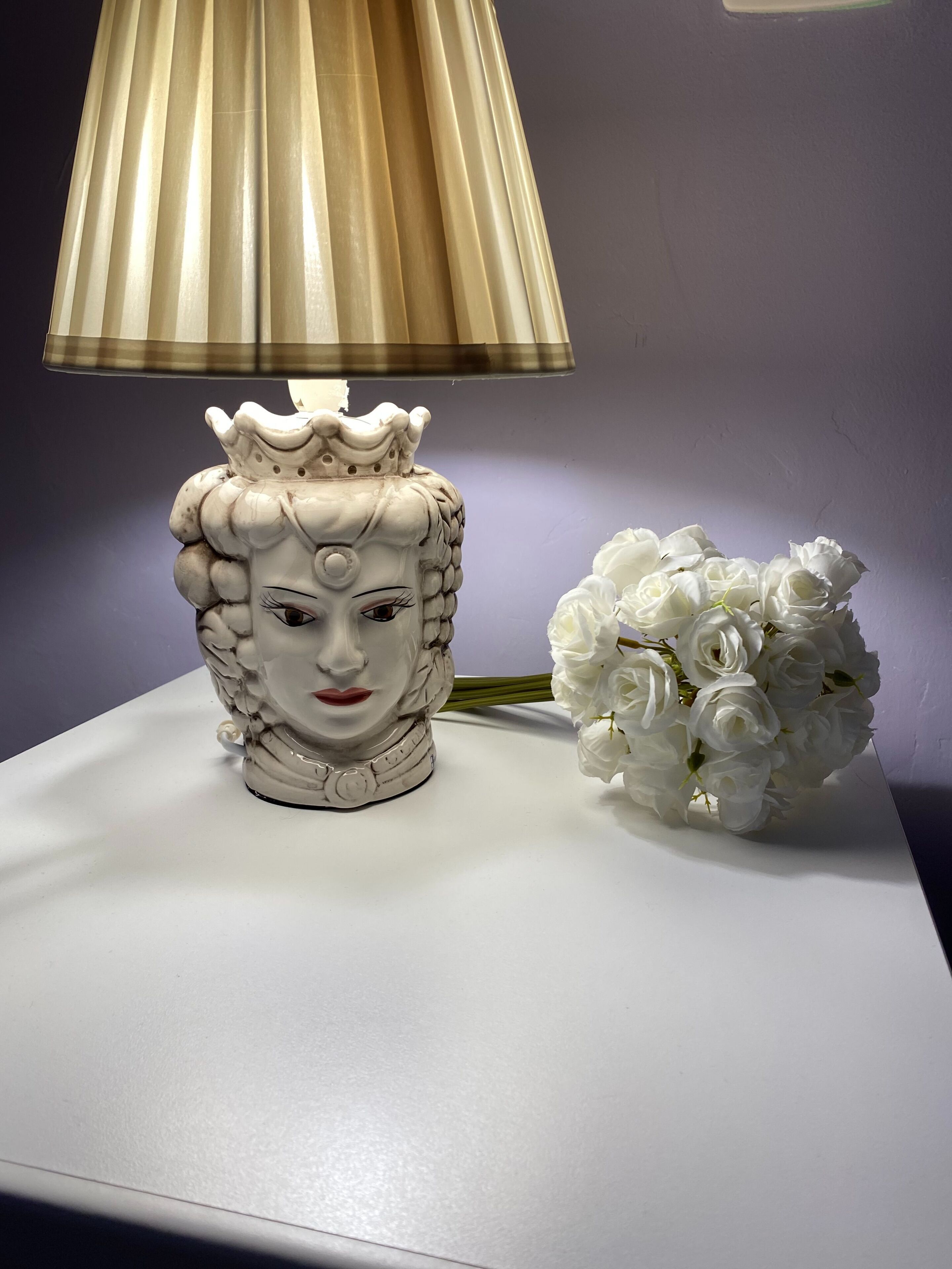 Una lámpara de cerámica blanca con diseño de corona y un arreglo floral blanco sobre una superficie blanca.