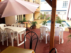 An outdoor patio with white plastic chairs and tables, a pink umbrella, and a view of a swimming pool and buildings in the background.