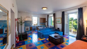 Un salon avec un canapé bleu, une table basse et un téléviseur. Au fond, on aperçoit une cuisine avec un coin repas.
