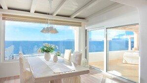 Un comedor moderno con vista al mar, una mesa blanca con un florero y una lámpara colgante.