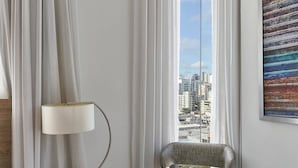 A modern room with a view of a cityscape through sheer curtains, a wooden nightstand with a lamp, and a grey upholstered chair. - Apartment N1001 Downtown Santo Domingo (Santo Domingo)