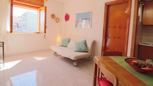 Une pièce comprenant un canapé, une table à manger avec un panier de fruits, une fenêtre avec des rideaux et une porte.