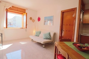 A room with a sofa, a dining table with a fruit bowl, a window with curtains, and a door. - Casa Myriam Cozy Two-room Apartment 3 Minutes From the Beaches of Otranto (Otranto)