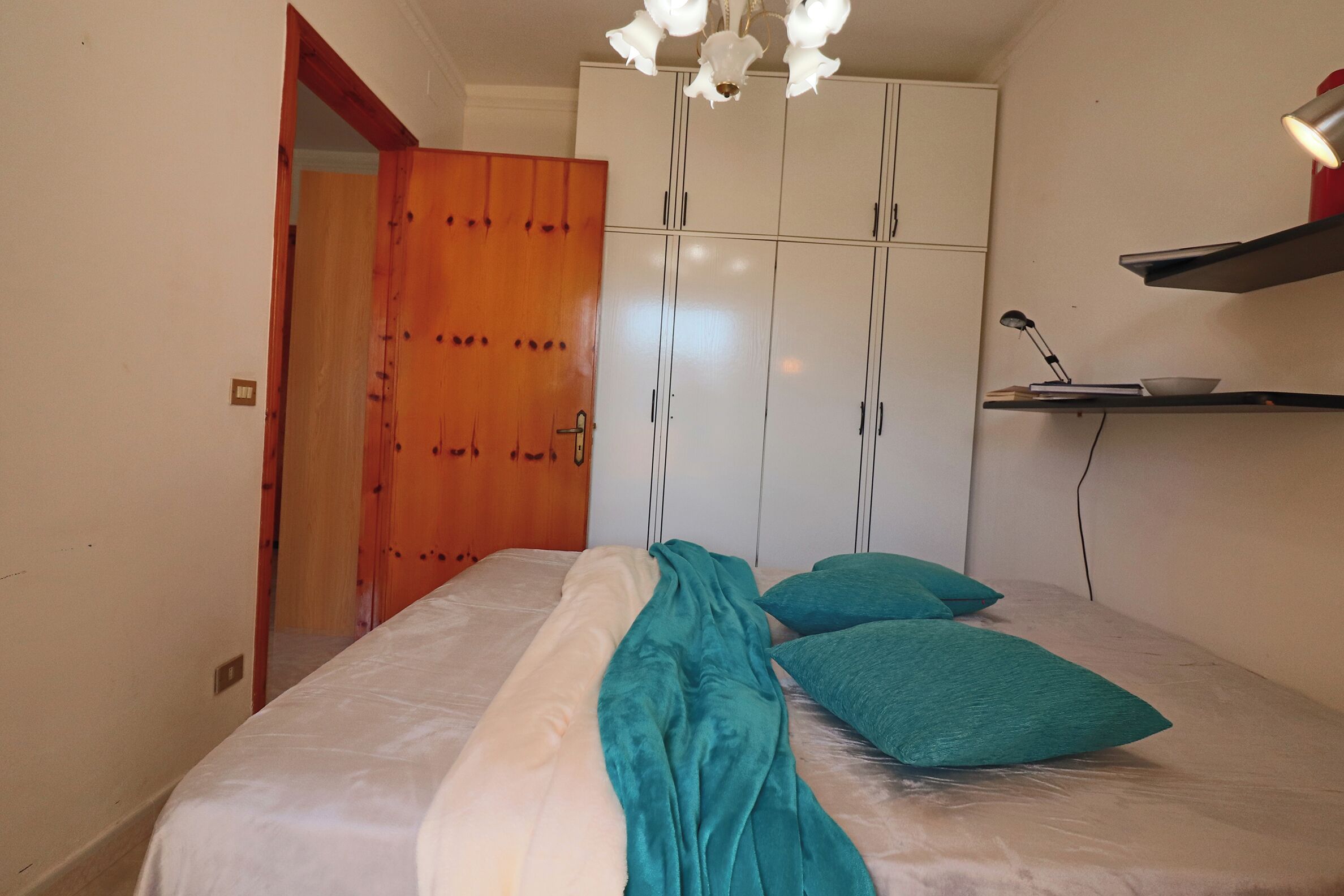 A bedroom with a bed, a wardrobe, a desk with a lamp, and a shelf with a bowl.