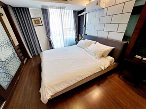 A neatly made bed in a hotel room with a headboard, wooden flooring, and a window with curtains.