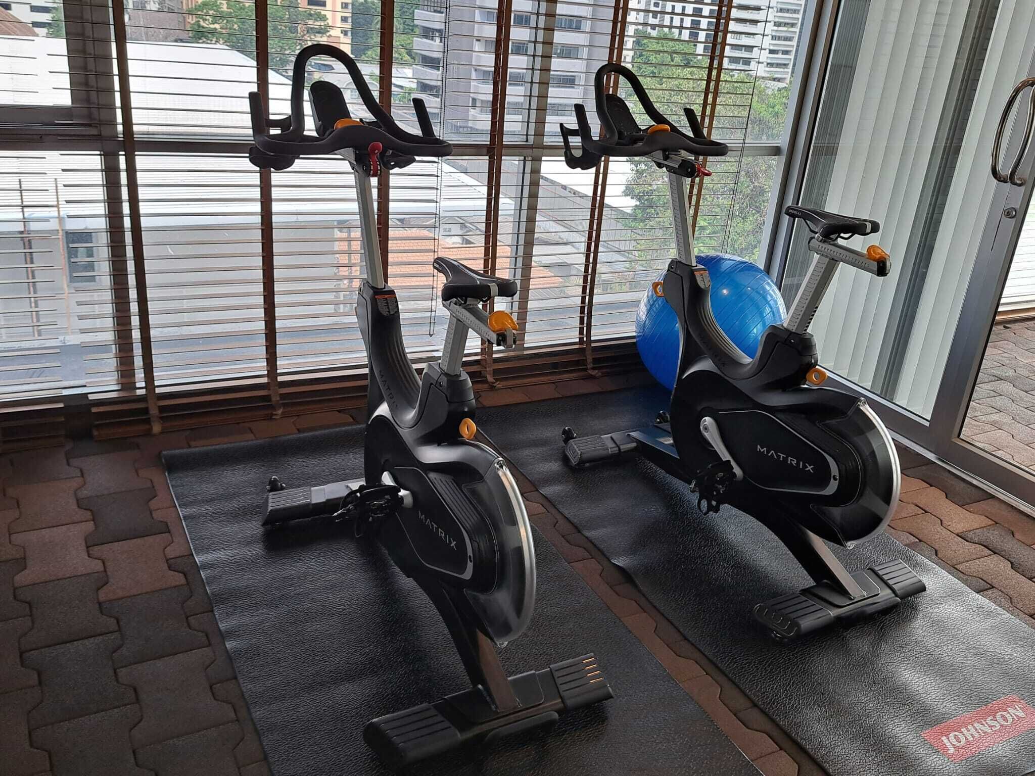 Dos bicicletas estáticas en un gimnasio con ventanas amplias y una pelota de ejercicios azul.