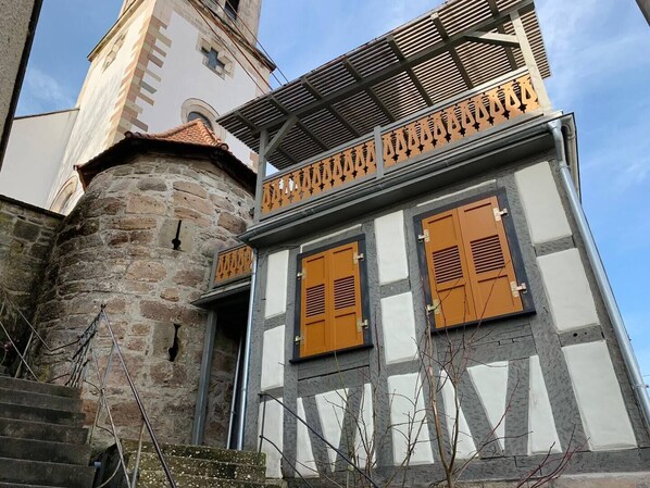 Un bâtiment historique doté d’une tour en pierre, de volets en bois et d’un balcon orné de ferronnerie ouvragée.