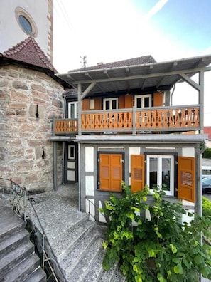 A two-story building with a stone facade, wooden shutters, and a balcony with a metal railing.