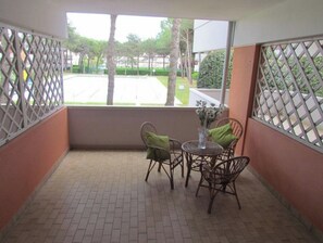 Varanda com cadeiras de vime e uma mesa, vista para a rua e árvores, e piso de azulejos.