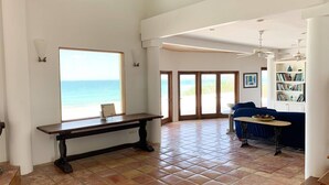 A spacious living area with a large window overlooking the ocean, a wooden bench, a blue sofa, and a bookshelf. - Private Beachfront Home (Great Harbour Cay)