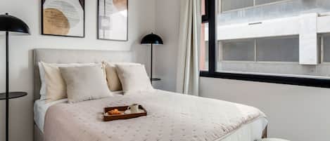 Quarto bem arrumado com cama, abajures, janela com vista para edifícios e obras de arte na parede.