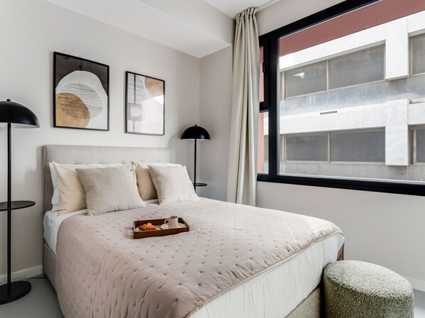A neatly arranged bedroom with a bed, bedside lamps, a window with a view of buildings, and artwork on the wall.