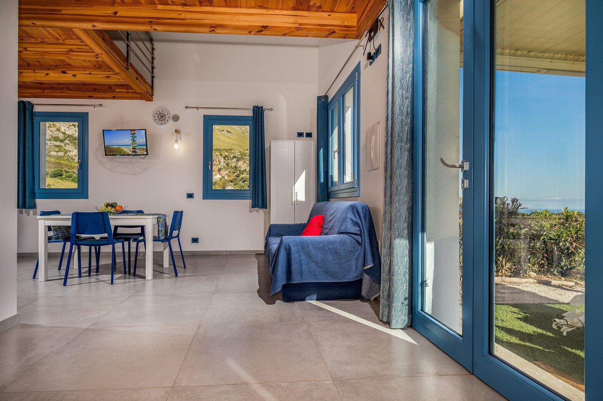 A spacious living area with a dining table, blue chairs, a sofa with a red pillow, and large windows offering a view of the outdoors.