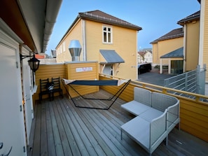 En träveranda med en hängmatta, ett bord och stolar, med utsikt över ett bostadsområde.