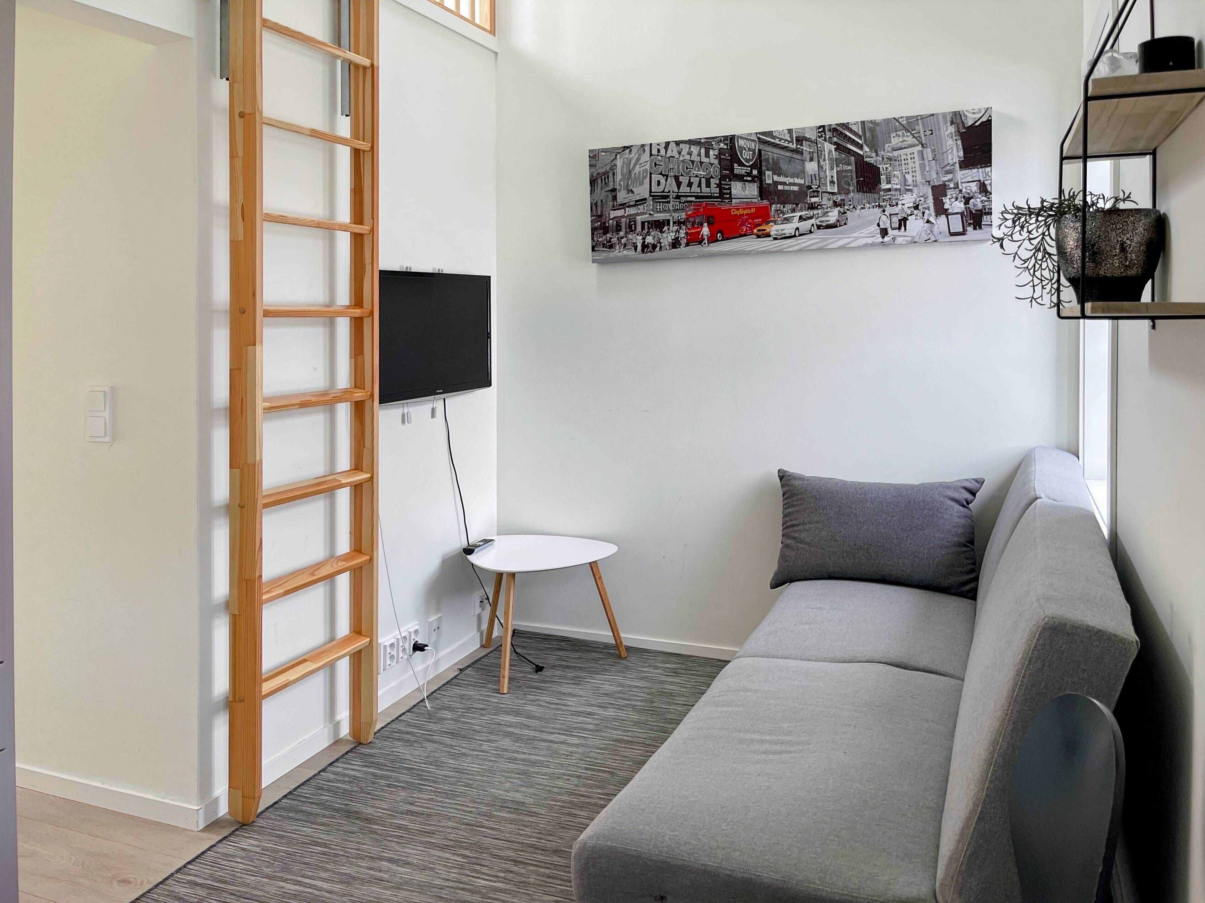 Un salon moderne avec un canapé gris, une petite table blanche, un téléviseur fixé au mur blanc et une échelle appuyée contre le mur.