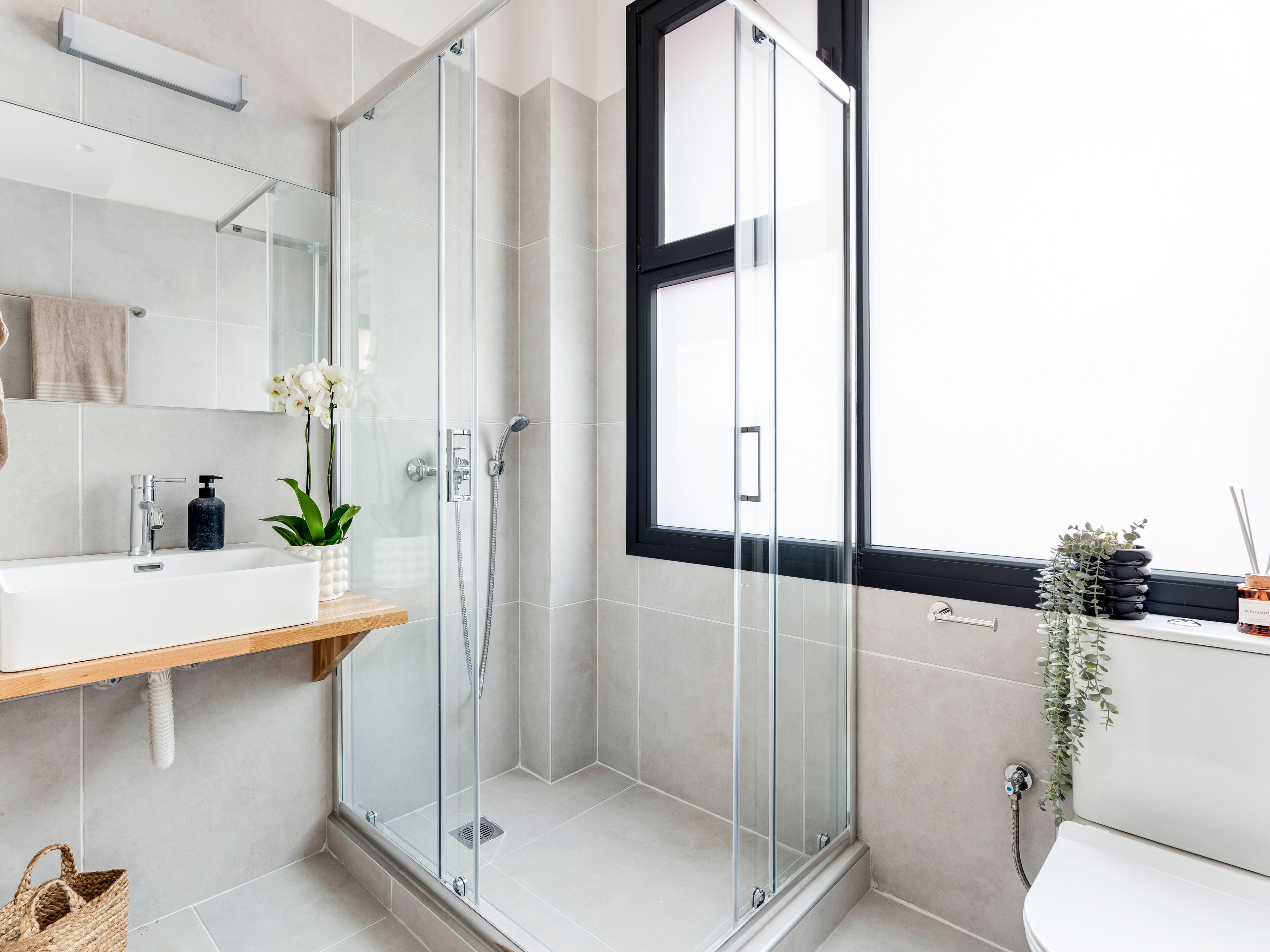 A modern bathroom with a glass-enclosed shower, a white sink with a wooden shelf, and a window providing natural light.