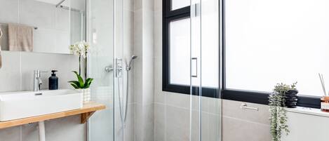 A modern bathroom with a glass-enclosed shower, a white sink with a wooden shelf, and a window providing natural light.