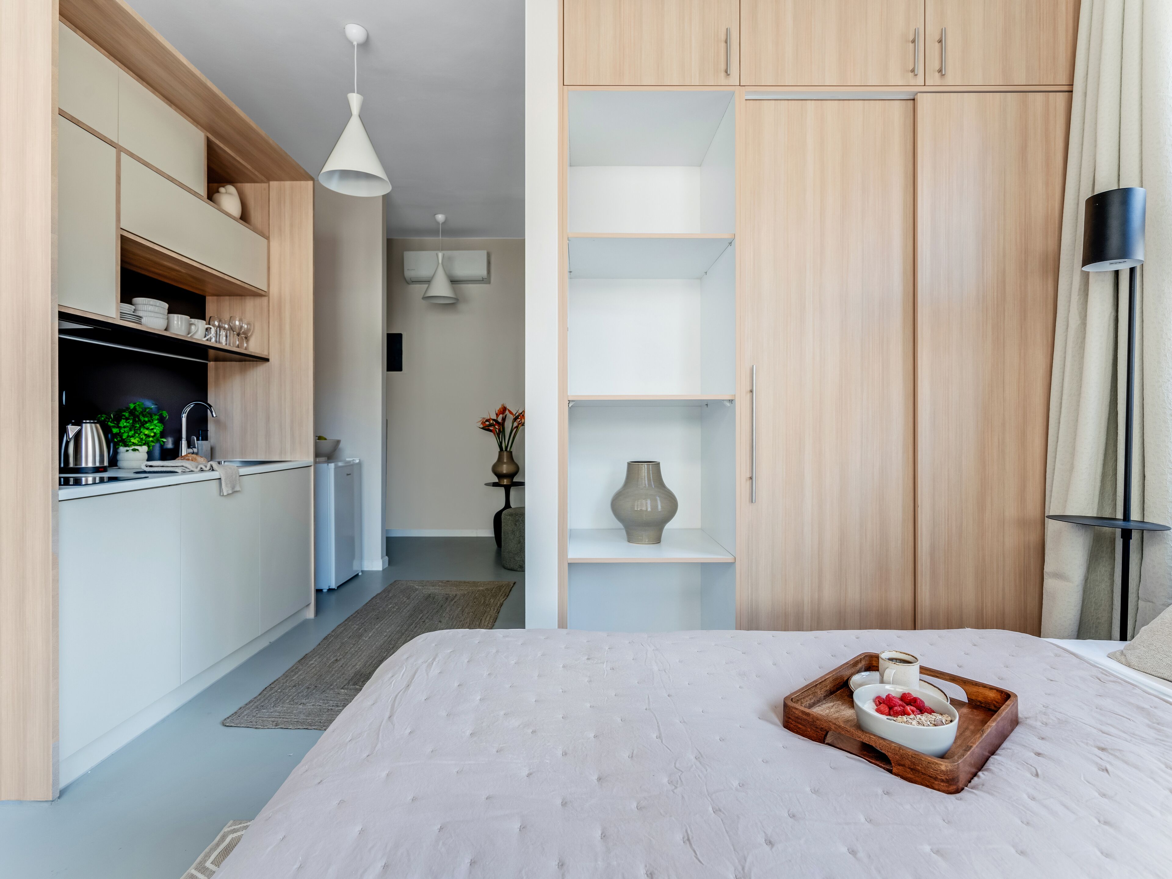 Quarto moderno com uma cama grande, mesa de cabeceira de madeira com bandeja de comida e uma kitchenette com armários e pia.