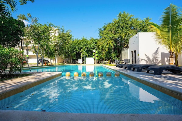 Une piscine à l’eau d’un bleu cristallin, entourée d’arbres et bordée d’un bâtiment blanc.
