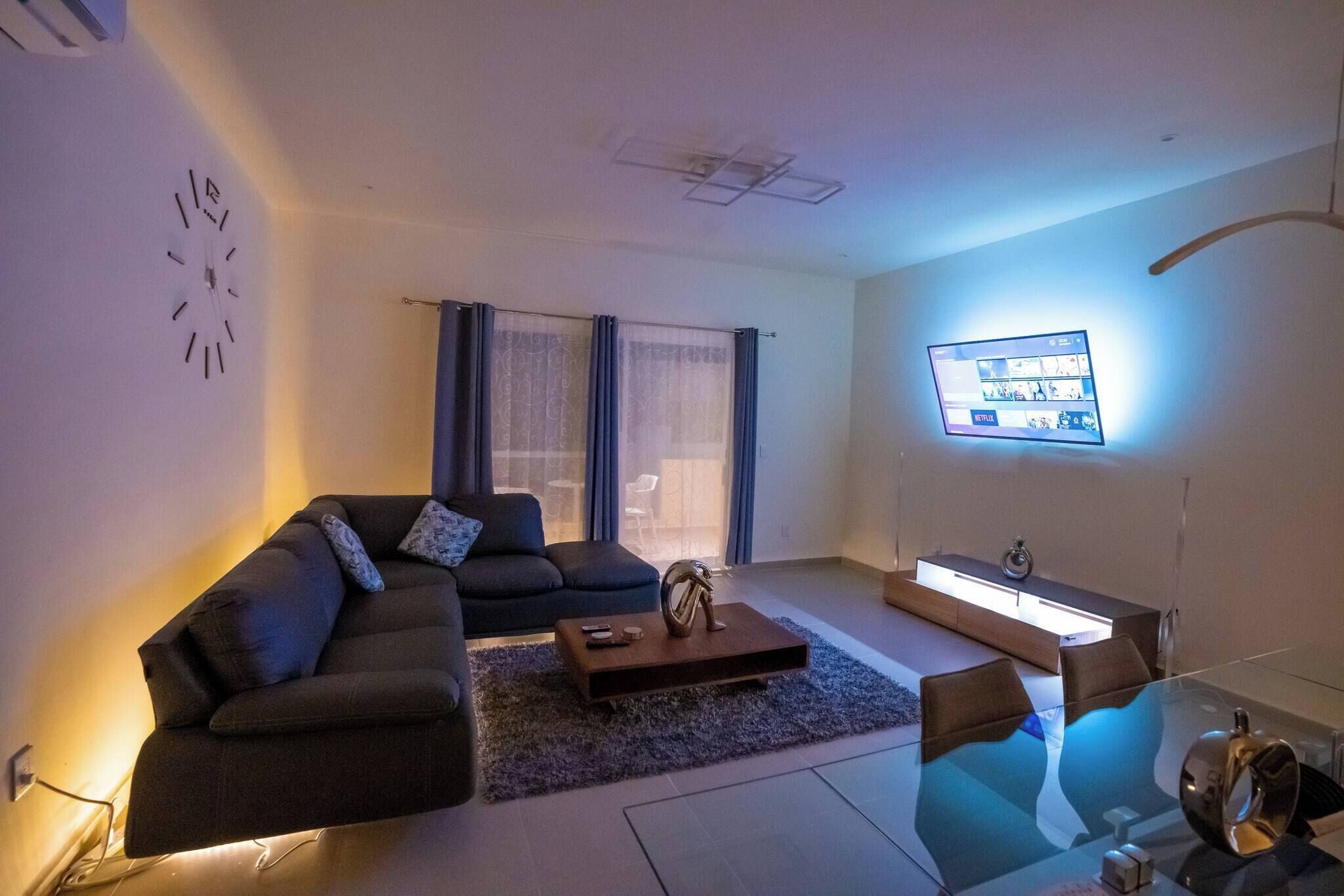 A modern living room with a sectional sofa, a glass coffee table, a TV mounted on the wall, and a sideboard with a light.