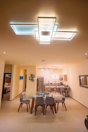 A modern kitchen with a central island, a dining area with a glass table, and a ceiling light fixture.