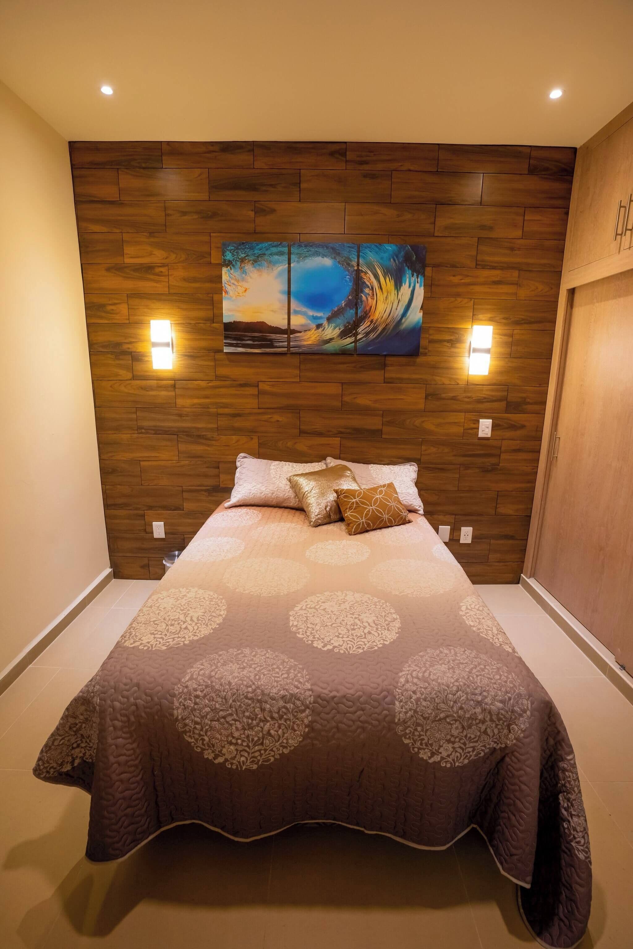 Une chambre à coucher avec une tête de lit en bois, un lit recouvert d’une couvre-lit à motifs, deux lampes fixées au mur et un tableau au-dessus du lit.