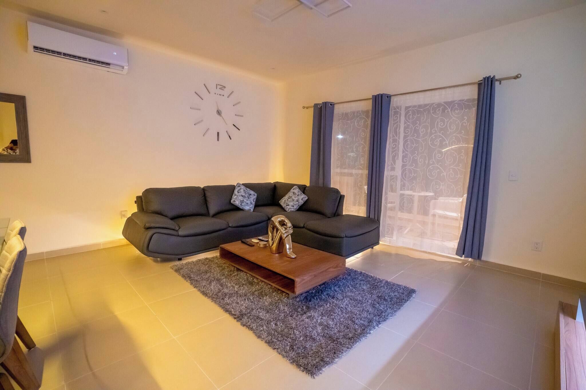 Una sala de estar con un sofá, una mesa de centro, un reloj y un ventanal con cortinas.