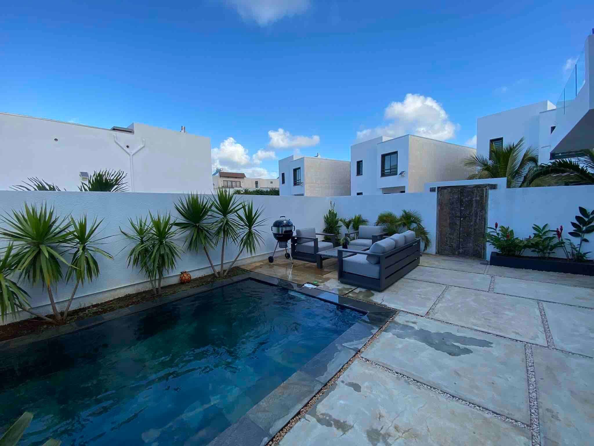 Un área de piscina exterior moderna con una parrilla incorporada, asientos y palmeras.