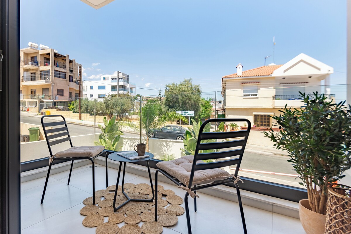 A balcony with two chairs and a small table, overlooking a street with buildings and a car.