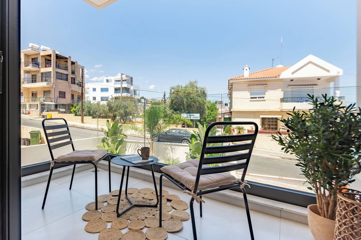 A balcony with two chairs and a small table, overlooking a street with buildings and a car.