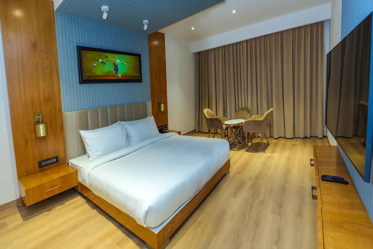 A hotel room with a large bed, a TV mounted on the wall, a desk with two chairs, and a large window with curtains.