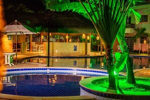 Un espace piscine nocturne doté d’une structure à toit de chaume, d’une petite piscine et d’un palmier.