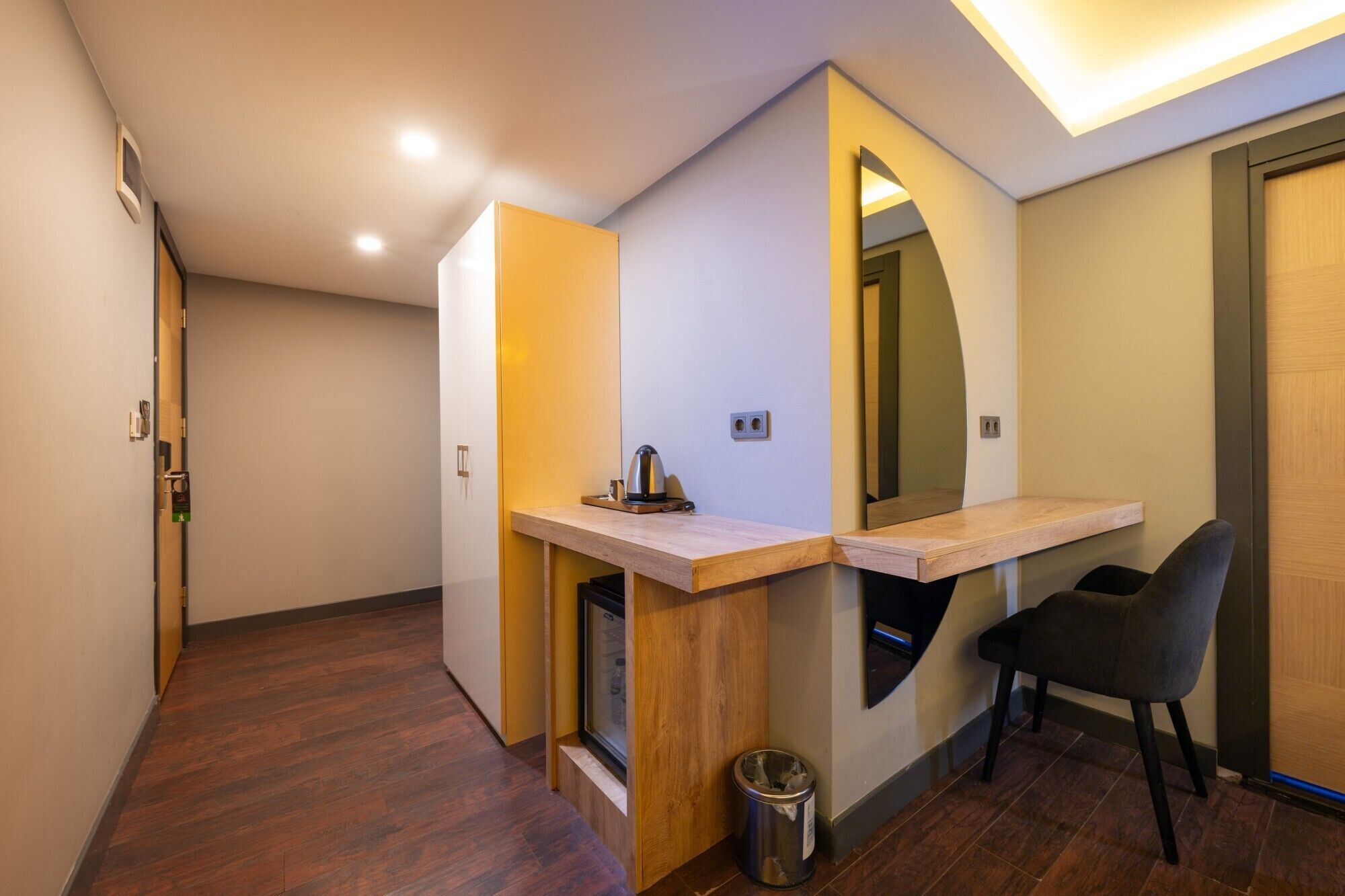 A modern hotel room with a wooden floor, a small desk with a kettle, a chair, and a trash bin.