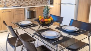 A modern kitchen with wooden cabinets, a tiled backsplash, and a glass dining table set with plates, cutlery, and a fruit basket. - Villa Cool with Private Pool (Malevizi)