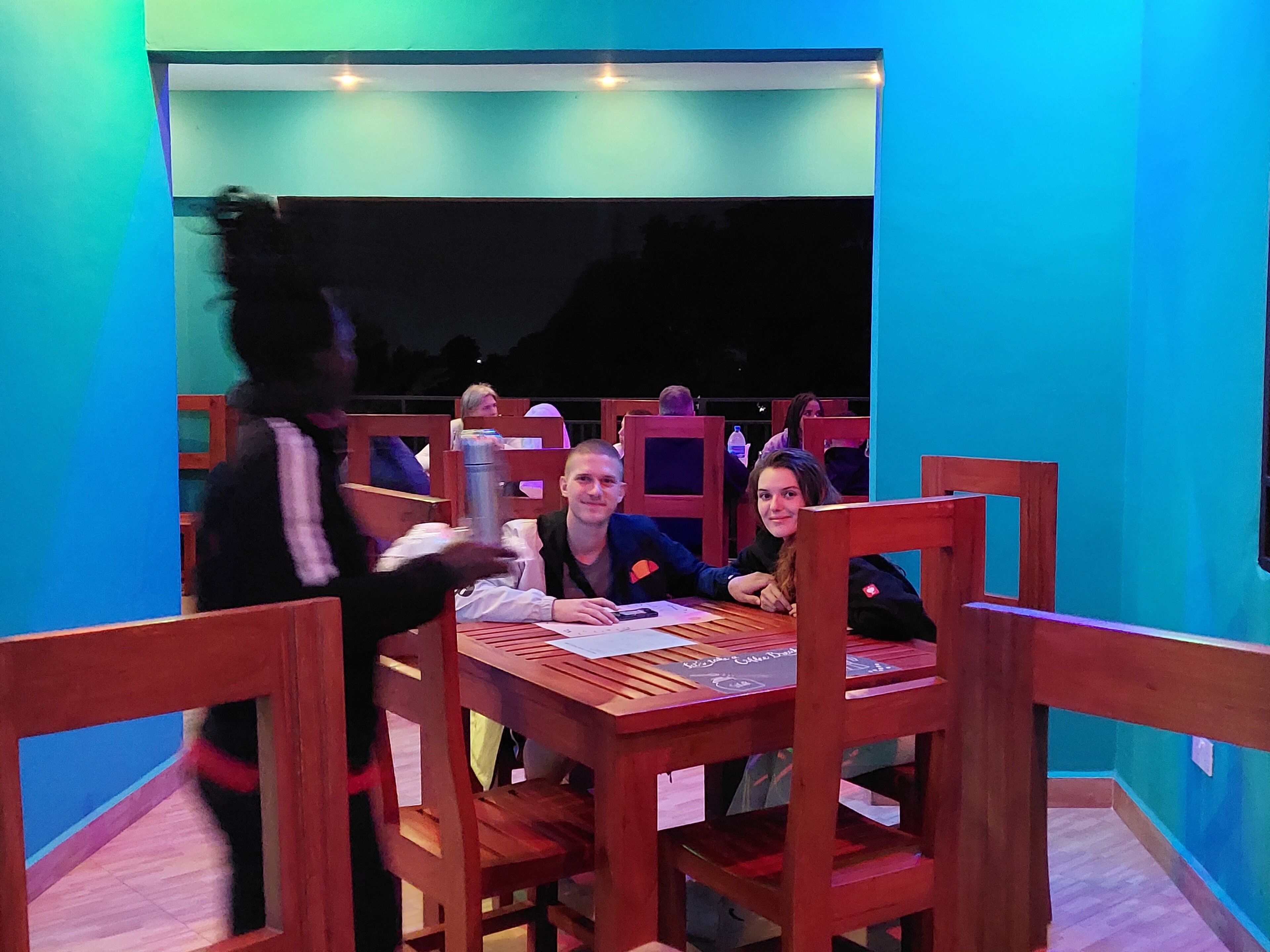 A restaurant with wooden tables and chairs, a blue interior, and a view of the night sky.