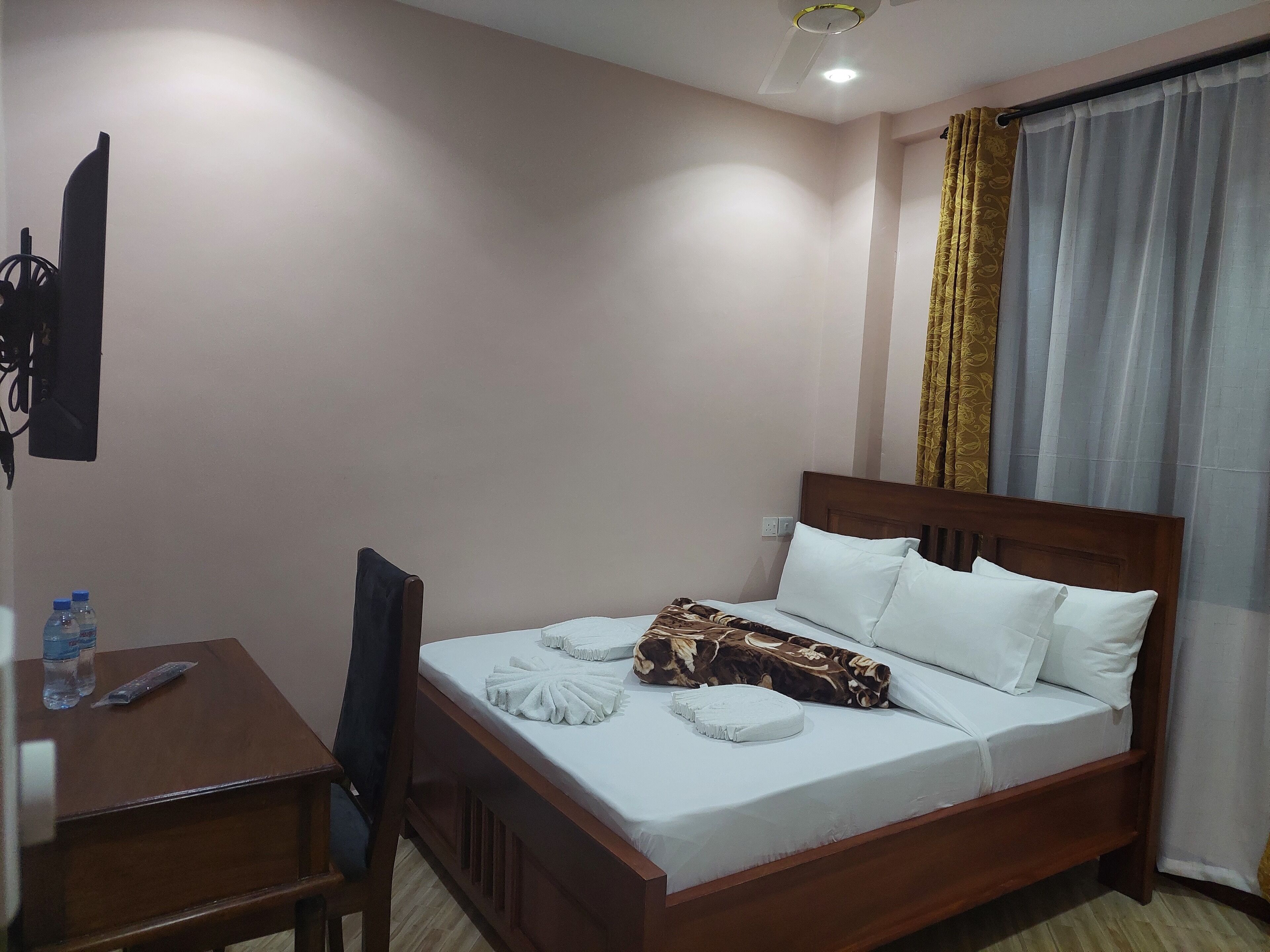 A neatly made bed with white linens and a brown patterned blanket, a wooden desk with a chair, and a water bottle on the desk.