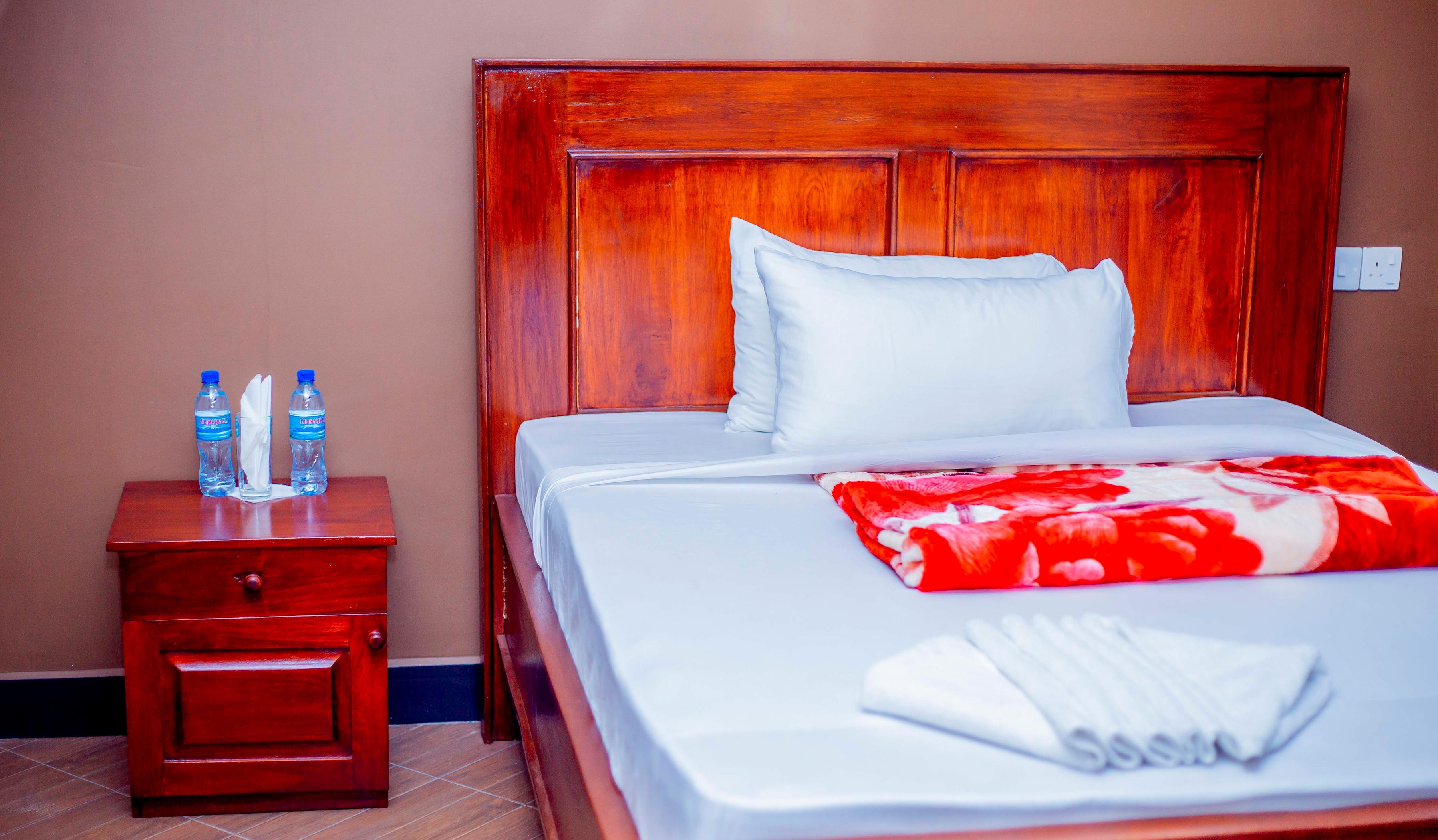 Ein Hotelzimmer mit einem hölzernen Kopfteil, einem ordentlich bezogenen Bett mit weißer Bettwäsche, einer roten und weißen Decke sowie einem Nachttisch mit zwei Wasserflaschen und einem gefalteten Handtuch.