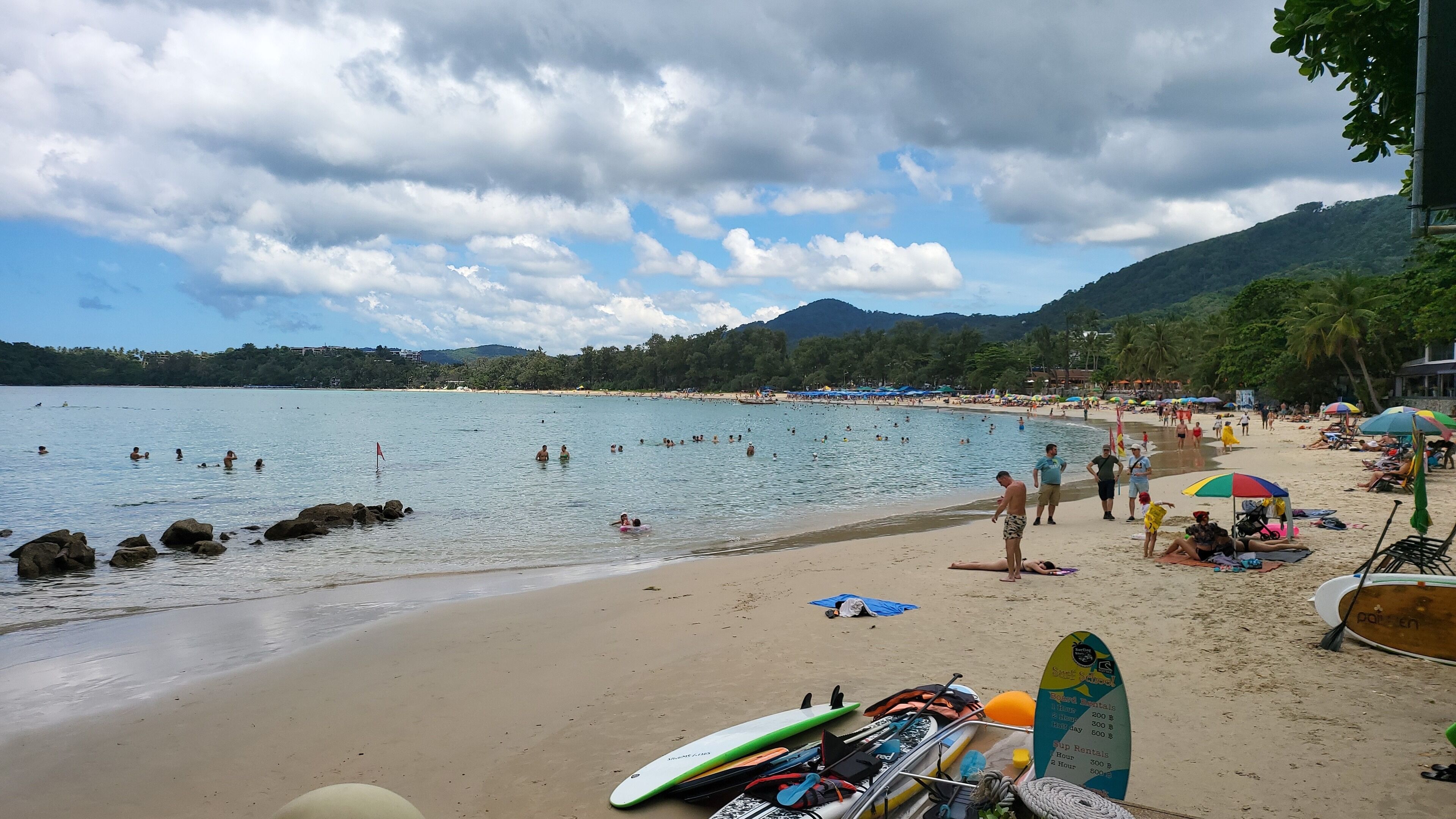 A beach with people swimming and sunbathing, a surfboard rack, and a sign for surf rentals.