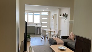 A modern living room with a sofa, coffee table, and TV stand. There is a kitchen in the background with a dining area.