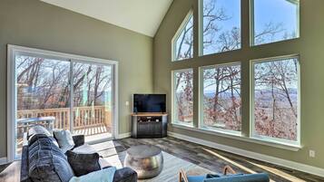 A spacious living room with a large TV, comfortable seating, and a balcony with a view of trees.